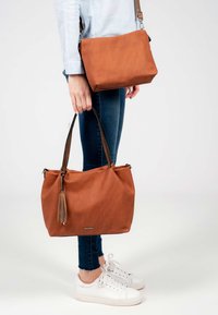Personne portant un jean bleu et des baskets blanches, tenant un grand sac fourre-tout marron et un plus petit sac bandoulière sur un fond uni.