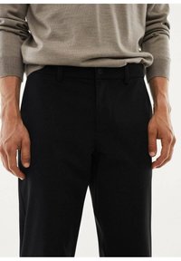 Pantaloni neri con una texture liscia e una vestibilità standard, dotati di chiusura a bottone e passanti per cintura, indossati con un maglione grigio chiaro.