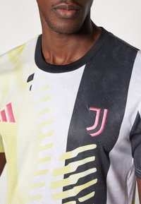 Maillot à manches courtes présentant des couleurs noir, blanc et lime avec un motif texturé, col rond, et un logo Juventus en rose bien visible.