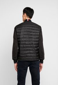 Zwarte pufferjack met een gewatteerd ontwerp, voorzien van een ribgebreide kraag met rode en witte strepen, en strakke mouwen. Draagt over donkere jeans.
