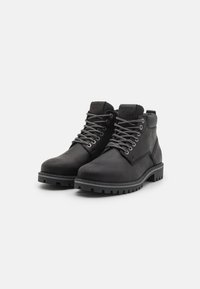 Par de botas de cuero negro con cordones de patrón gris, cuellos acolchados y suelas de goma gruesa sobre un fondo blanco.