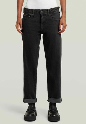 Bas du corps d'une personne portant un jean droit noir à revers, des mocassins noirs et un haut blanc sur un fond clair uni.
