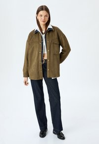 Camicia overshirt in suede verde oliva con colletto in denim bianco, tasca frontale e bottoni a pressione, abbinata a pantaloni in denim scuro e scarpe nere.
