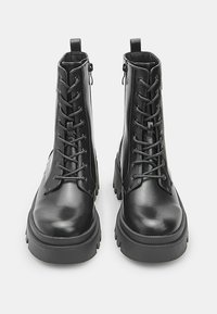 Stivali da combattimento neri in pelle con lacci, suole spesse e cerniere laterali, visti frontalmente su uno sfondo semplice.