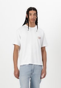 White cotton t-shirt with short sleeves, relaxed fit, featuring small red embroidered text on the left chest. Paired with light blue jeans.