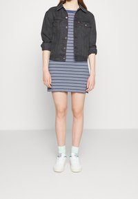 Veste en denim noire portée sur une robe rayée bleu et blanc. Des baskets blanches et des chaussettes bleu clair complètent la tenue. Design décontracté et ajusté.