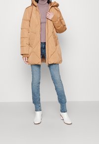 Béžová vatovaná bunda s kapucí, diagonálním prošíváním a předním zipem. Doplněná světle modrými džíny a bílými kotníkovými botami.