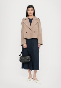 Veste beige courte avec double boutons, associée à un haut bleu marine et un pantalon large. Sac à main matelassé noir et chaussures pointues blanches.