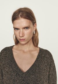 Kobieta z prostymi, jasnobrązowymi włosami nosi srebrne kolczyki w kształcie koła oraz ciemny sweter z teksturowanego dzianiny z głębokim dekoltem w kształcie litery V, na jednolitym tle.