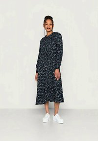 Vestido preto floral com mangas longas, cintura franzida e comprimento midi, apresentando pequenos padrões de flores multicoloridas. Combinado com ténis brancos.