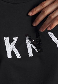 Svart tygplagg med bokstäverna "KNY" gjorda av reflekterande paljetter. Paljetterna växlar mellan vitt och svart. En hand är synlig.