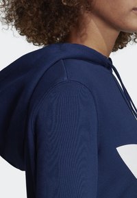 Navy sininen huppari, jossa on suuri valkoinen logo, ribbaustuotteet hartioissa ja nyörit kaulassa. Pehmeä tekstuuri, rento muotoilu.