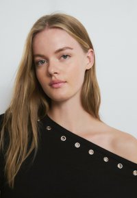 Mujer joven con cabello largo y rubio, vestida con un top negro de hombros descubiertos con detalles de ojales plateados, sobre un fondo claro y liso.