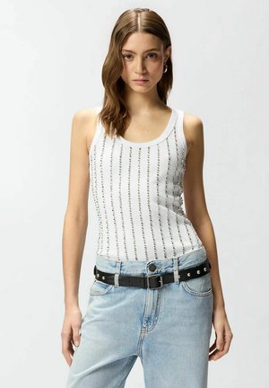 Donna con capelli castani a lunghezza spalle che indossa una canotta bianca con strisce verticali argento e jeans azzurri chiari con una cintura nera con borchie.