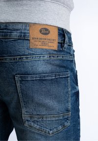 Džíny z denimu v tmavě modrém odstínu s koženou značkovou záplatou označenou nápisem „Petrol“, se dvěma zadními kapsami a šitím detailů.