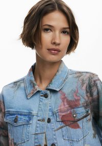 Veste en denim bleu présentant des motifs floraux dans des teintes de rose et de vert, avec des poches avant et des boutons-pression en métal. Design court avec col.