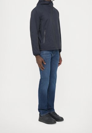 Homme portant une veste à capuche noire zippée, un jean bleu et des baskets noires, debout dans une pose neutre sur un fond blanc.
