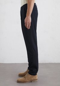 Pantalon bleu marine en tissu lisse, avec une coupe slim et une poche latérale discrète. Assorti à des chaussures en daim marron clair.