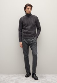 Grauer Rollkragenpullover aus Kabelstrick kombiniert mit grauen gestreiften Hosen und schwarzen Slippern. Minimalistische Gestaltung vor neutralem Hintergrund.