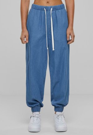 Personne portant un pantalon de jogging bleu ample avec un cordon blanc et des poignets élastiques, associée à des baskets blanches, debout devant un fond uni.