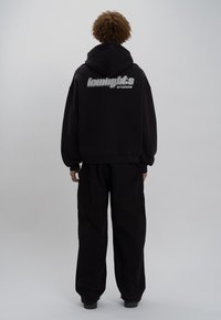 Felpa nera con cappuccio e logo grafico sulla schiena, abbinata a pantaloni neri larghi, indossata da un modello di spalle, su uno sfondo chiaro.