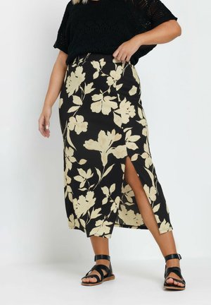 Larga falda negra con un estampado floral en color crema, que presenta una abertura lateral. Combinada con sandalias negras de tiras y una blusa negra holgada.