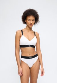 Γυναίκα που φοράει λευκό σουτιέν και λευκά εσώρουχα με μαύρες ελαστικές ταινίες και λογότυπο Calvin Klein, στέκεται μπροστά σε απλό λευκό φόντο.