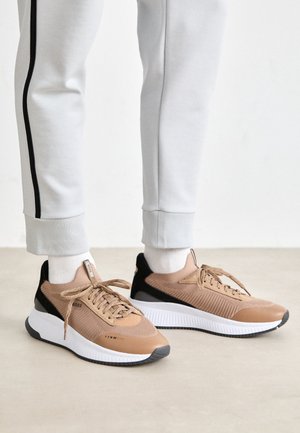 Person trägt beige und schwarze Sneakers mit weißen Sohlen und hellgraue Hose mit einem schwarzen Streifen und steht auf einem hellen Boden.