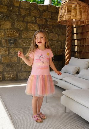 Lächelndes Mädchen mit langen Haaren, das ein pinkes Einhorn-T-Shirt und einen bunten Tüllrock trägt, steht drinnen an einer Steinwand und einem weißen Sofa.