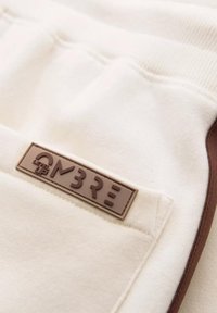 Pantalones deportivos beige con una textura suave, que presentan un parche de logo de goma marrón y acentos laterales marrones en contraste.