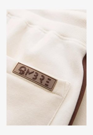 Pantalons de survêtement beige avec une texture lisse, ornés d'un patch logo en caoutchouc marron et d'accents latéraux contrastants en marron.