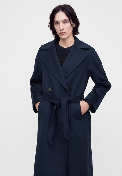 Personne aux cheveux courts et foncés portant un manteau bleu marine foncé ceinturé par-dessus un pull noir, debout les mains dans les poches du manteau devant un fond uni.