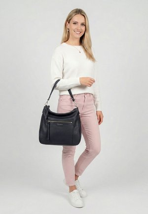 Mujer con un suéter blanco y pantalones rosas, de pie descalza con un bolso de cuero negro colgado del brazo izquierdo.