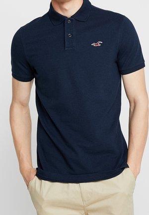 Mann trägt ein navyblaues kurzärmliges Poloshirt mit einem kleinen rot-weißen Logo auf der Brust, beige Hose, Hände in den Taschen.