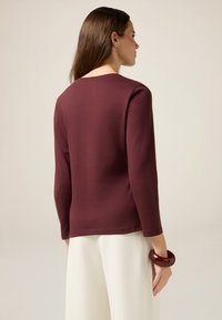 Bordeauxrotes Langarmoberteil aus glattem Stoff, mit einem runden Halsausschnitt und geradem Saum. Kombiniert mit hellen, weiten Hosen und einem dunklen Armreif.