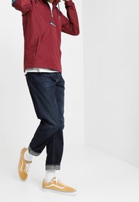 Chaqueta ligera roja con media cremallera, acentos azules, junto a unos vaqueros azul oscuro y zapatillas amarillas, llevada con calcetines blancos.