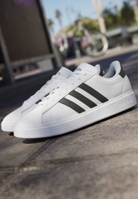 Weiße Adidas Sneakers mit schwarzen Streifen, draußen auf Betonpflaster mit verschwommenem Fahrrad und Bäumen im Hintergrund.