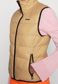 Hnědá péřová vesta s černým zipem a lemem obsahuje logo. Je vrstvená přes světle růžový svetr s rozšířenými rukávy.