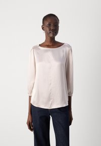 Light pink blouse with three-quarter sleeves, round neckline, smooth texture, and subtle seam detail, paired with dark blue jeans.