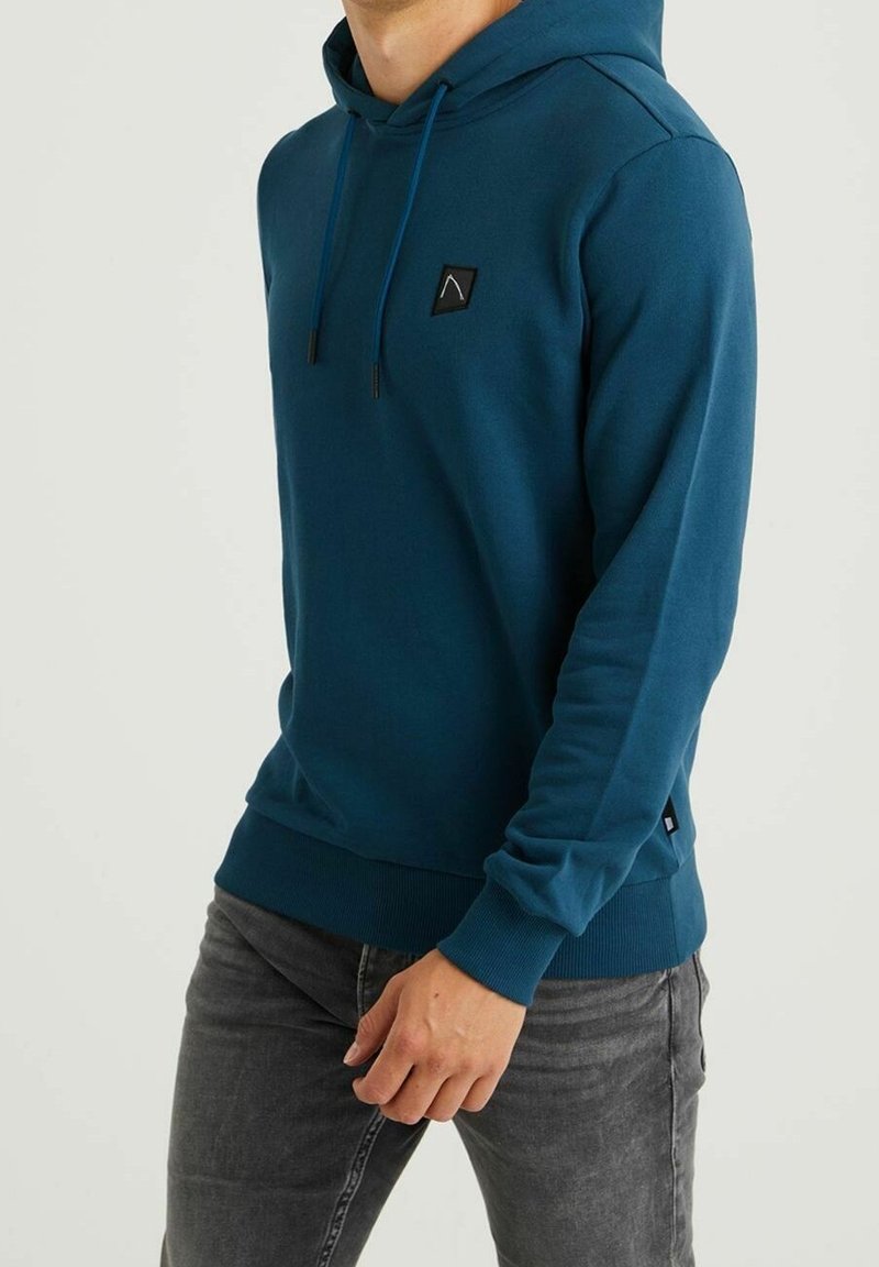 Homme portant un sweat à capuche bleu sarcelle avec un petit patch noir sur la poitrine et un jean gris foncé, debout devant un fond uni.