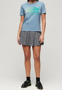 Ljusblå bomull T-shirt med grön grafisk text, kombinerad med en svart och vit rutigt plisserad kjol. Svarta skor och strumpor fulländar utseendet.