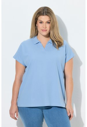 Kobieta o blond włosach, nosząca srebrne kolczyki-koła, jasnoniebieską koszulkę polo z krótkim rękawem i niebieskie dżinsy na białym tle.