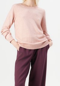Pull rose clair en matière douce avec un col rond et un ourlet côtelé, associé à un pantalon ample de couleur violet foncé.