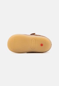 Suela de zapato de ante marrón con una parte inferior de goma amarilla texturizada. Presenta el nombre de la marca "Kickers" y un punto rojo para mayor tracción.
