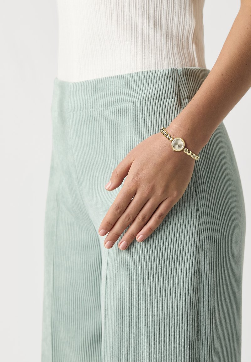Pohodlné ribbed nohavice v mentolovej zelenej farbe sú skombinované s bielym priliehavým topom. Na zápästí sa nachádza zlatý náramok s okrúhlou fasádou a korálkovými akcentmi.