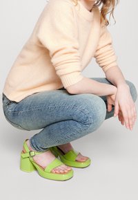 Zelené sluneční podpatky s lesklým povrchem, které mají široký pásek přes prsty a blokový podpatek pro stabilitu.