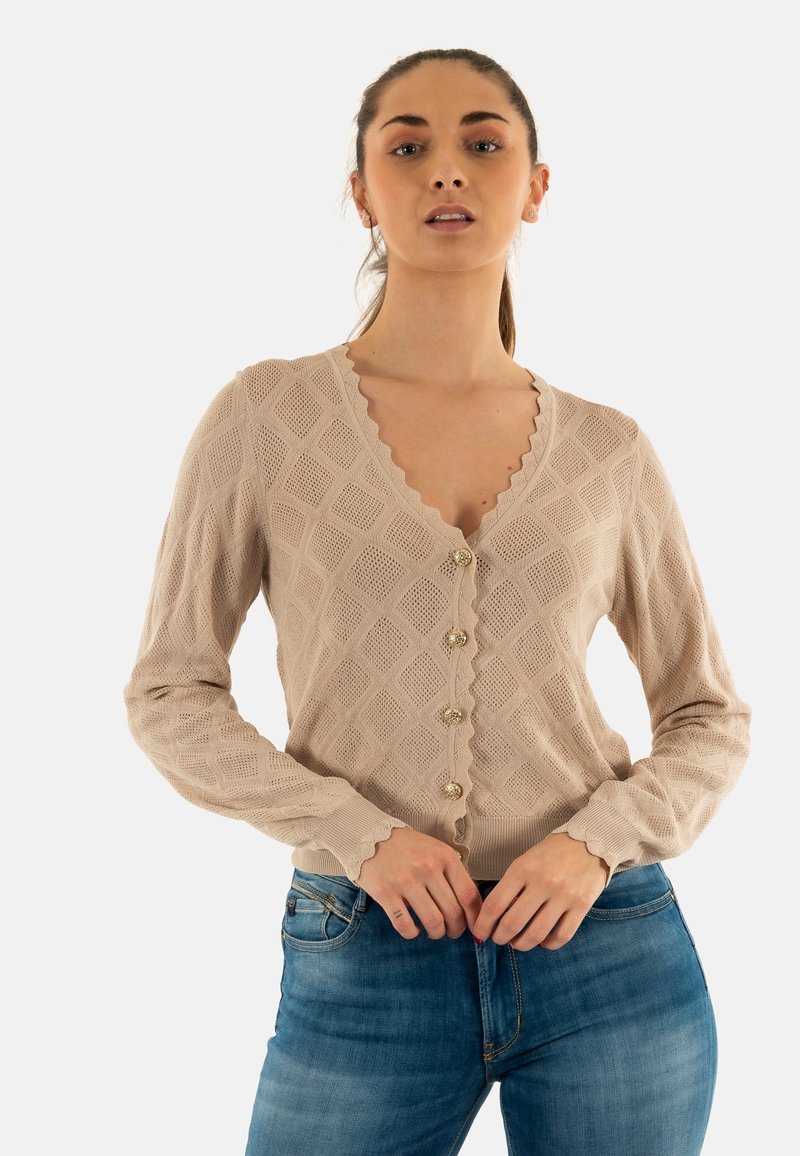 Cárdigan de punto beige con un patrón de diamantes, cuello con bordes en forma de ola y cierre de botones. Mangas largas con puños acanalados. Se lleva con vaqueros azules.