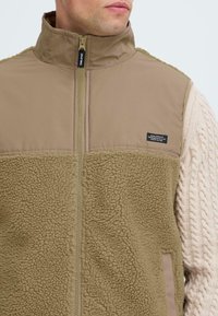 Brown zip-up vest featuring a fleece lower section, smooth upper fabric, and a small logo on the chest; textured and soft.