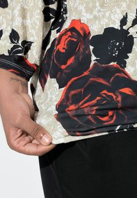Tričko s květinovým vzorem, červenými a černými růžemi na béžovém pozadí, s krátkými rukávy a volnějším střihem, doplněné černými kalhotami.