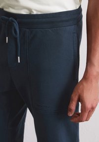 Marineblaue Sweatpants mit einem Kordelzugbund, ausgestattet mit seitlichen Taschen und einer strukturierten Oberfläche. Logo auf dem Bein geprägt.
